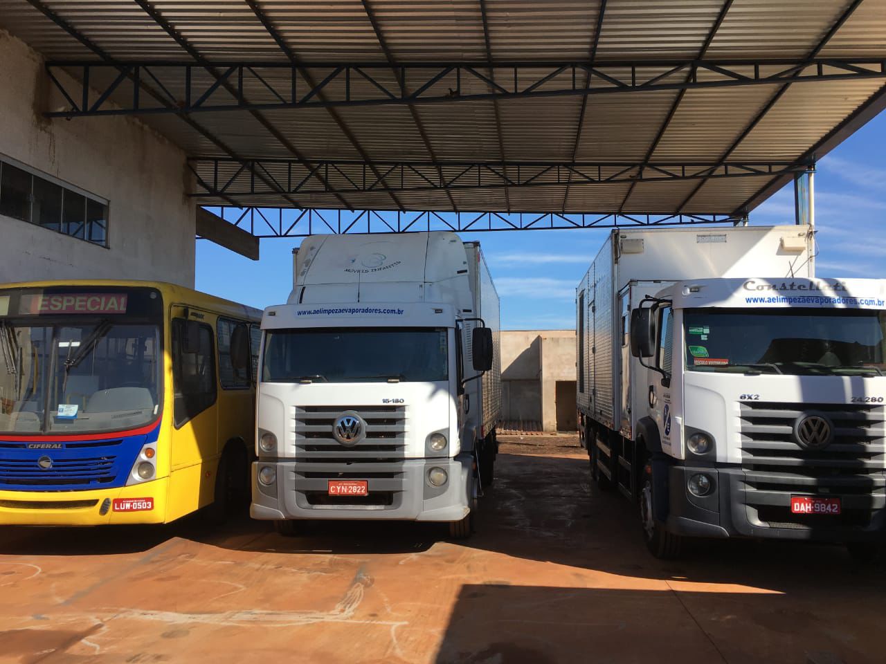Dois caminhões de carga grandes e um ônibus amarelo estacionados lado a lado sob uma garagem coberta, representando uma frota de serviços pesados.