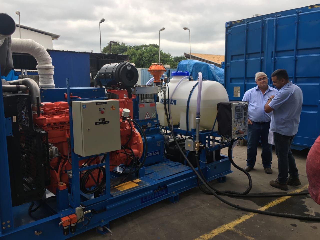 Dois homens de camisa azul inspecionando uma grande máquina de limpeza industrial com bombas, tubulações e tanques em uma instalação ao ar livre.