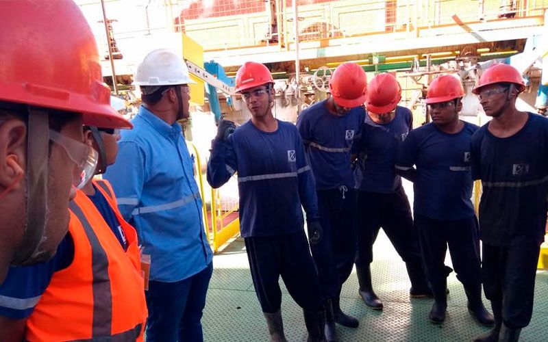 Grupo de funcionários em uniformes azuis e laranja com capacetes de segurança vermelhos reunidos durante uma reunião de segurança ou operacional em uma instalação industrial.