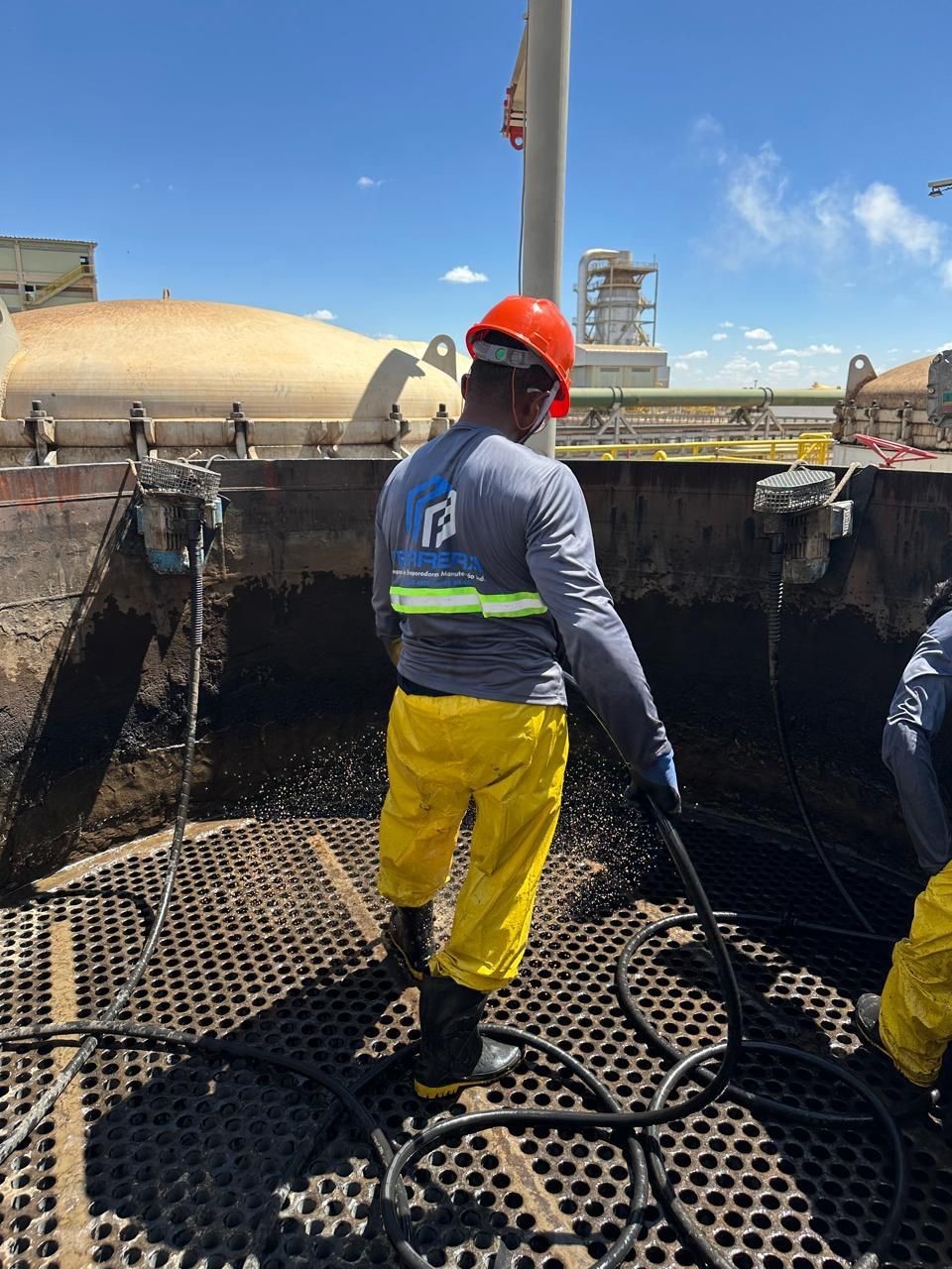 Funcionário usando capacete vermelho, camisa cinza com o logotipo da empresa, calças impermeáveis ​​amarelas e botas usando uma mangueira de alta pressão dentro de um grande tanque de evaporador industrial.