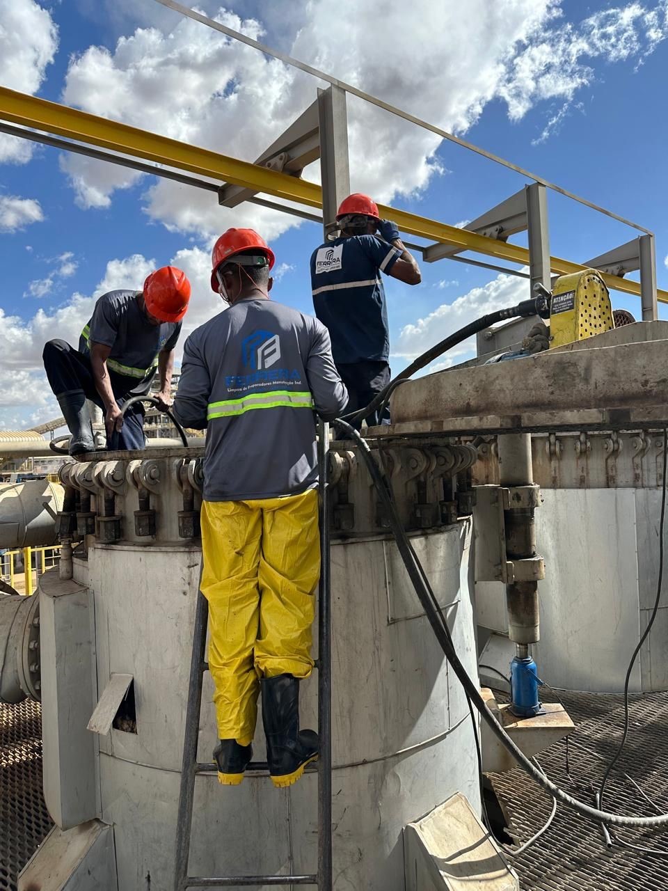 Trabalhadores com capacetes de segurança e uniformes realizando manutenção em cima de um evaporador, um em pé em uma escada com mangueira enquanto os outros auxiliam, contra um fundo de céu nublado.