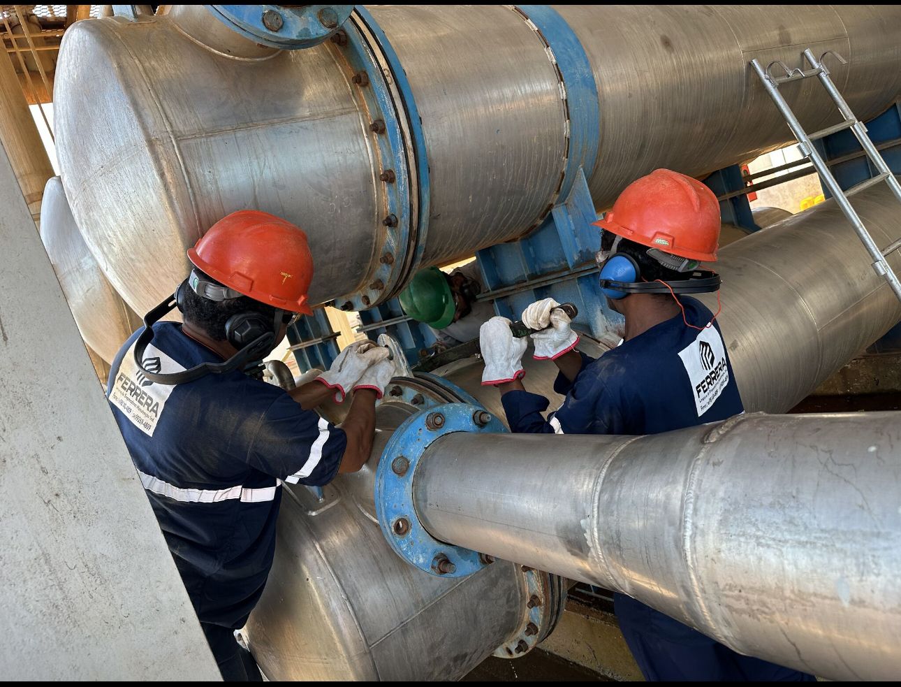 Dois profissionais da Ferrera trabalhando na manutenção de uma flange ou junta de tubulação de grande diâmetro em uma instalação industrial.